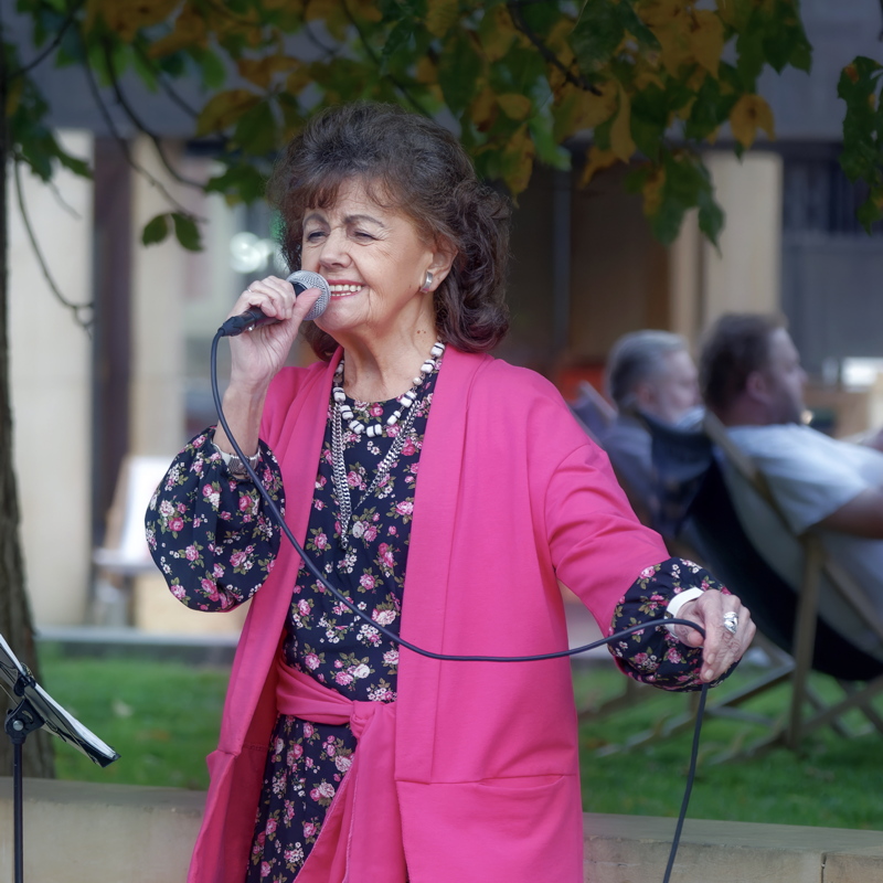 16 - Woman singing into a microphone while wearing a bright pink jacket and a floral dress